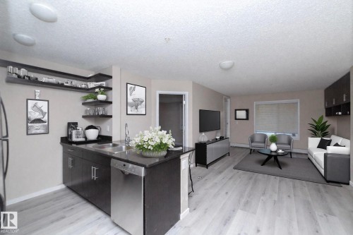 Open-concept living area featuring wood-finish flooring, a kitchen island with integrated stainless steel dishwasher, and dark cabinetry with open shelving - 108 530 Watt Boulevard, Edmonton, AB 