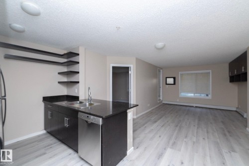 Open-concept kitchen and living area featuring dark wood cabinetry, a stainless steel dishwasher, a kitchen island with an integrated sink, dark countertops, and wood-finish flooring - 108 530 Watt Boulevard, Edmonton, AB 