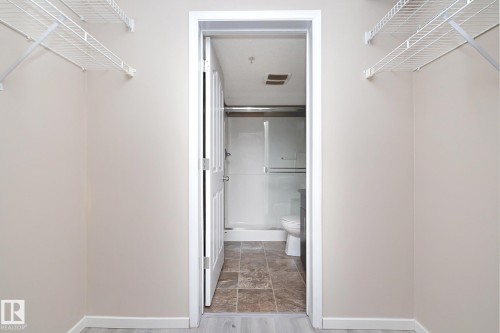 Walk-in closet featuring wire shelving, light-toned wall paint, and white trim, with a bathroom beyond the doorway showcasing a toilet, shower with sliding glass doors, and tiled flooring - 108 530 Watt Boulevard, Edmonton, AB 