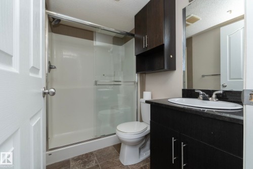 Bathroom featuring a dark wood-finish vanity with a dark countertop and integrated sink, a full-width mirror, a shower with sliding glass doors, and tile flooring - 108 530 Watt Boulevard, Edmonton, AB 