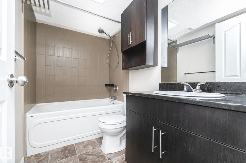 Bathroom featuring a white bathtub, beige tiled shower surround, dark wood-finish vanity with an integrated sink, dark countertop, and tiled flooring - 108 530 Watt Boulevard, Edmonton, AB 
