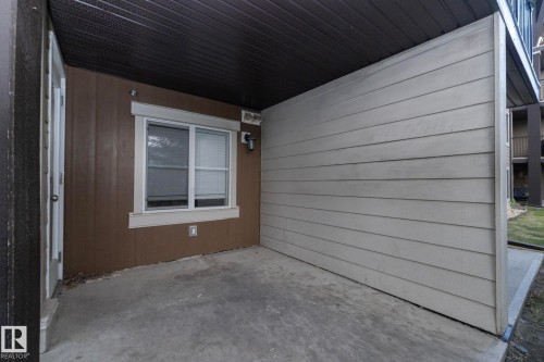 Covered patio area featuring a concrete slab, brown vertical siding, and a white-framed window with blinds - 108 530 Watt Boulevard, Edmonton, AB 