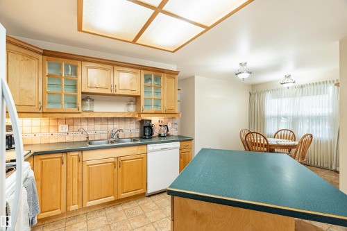 Kitchen featuring light wood cabinetry with frosted glass inserts, a double basin sink, and a decorative tile backsplash - 9011 60 Street, Edmonton, AB 