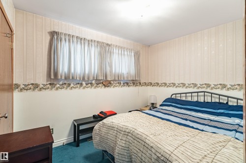 Carpeted room featuring a window with drapes, patterned wallpaper, a light wood-finish door, and a ceiling-mounted light fixture - 9011 60 Street, Edmonton, AB 