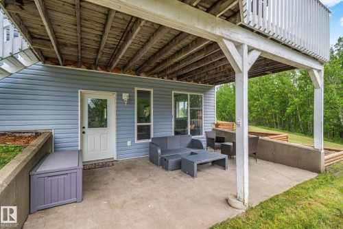Covered concrete patio featuring a white exterior door with glass insert, vertical siding, and an overhead wood-plank deck structure - 14 53122 Rge Road 14, Rural Parkland County, AB 