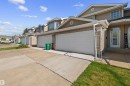 Townhome exterior featuring tan siding, brick accents, white garage doors, a concrete driveway, and a small front lawn - 5 20 Deerbourne Drive, St. Albert, AB 