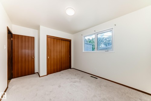 Carpeted room with white walls, featuring a standard window, a flush-mount ceiling light fixture, a wood-paneled hinged door, and a wood-paneled sliding closet door - 12022 62 Street, Edmonton, AB 