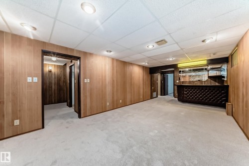 Spacious room featuring light-toned wood paneling, light carpet flooring, recessed lighting, and a built-in bar with a tufted dark facade - 12022 62 Street, Edmonton, AB 