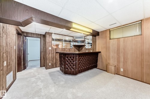 Finished basement area featuring a built-in bar with tufted leather-look paneling, wood-paneled walls, light-colored carpeting, and a drop ceiling with recessed lighting - 12022 62 Street, Edmonton, AB 