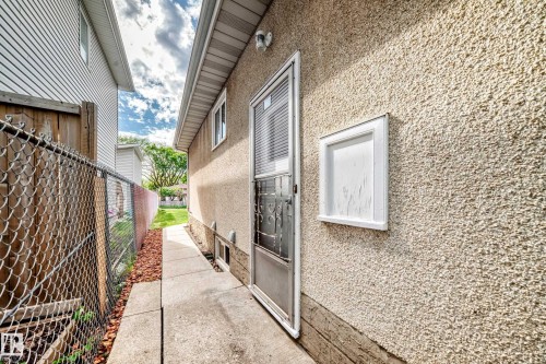 Stucco exterior featuring a side entrance door with screen, a basement window, and a concrete pathway - 12022 62 Street, Edmonton, AB 