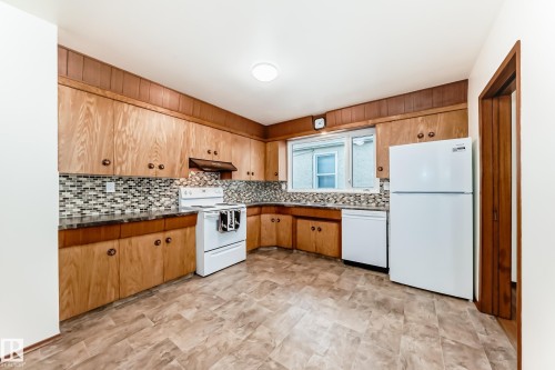 Kitchen featuring extensive wood cabinetry, coordinating wood paneling, white appliances, dark countertops, and a patterned tile backsplash - 12022 62 Street, Edmonton, AB 