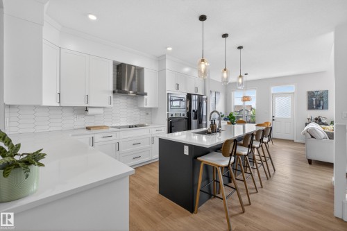 Modern kitchen featuring white cabinetry, a dark-toned island with seating, a stainless steel range hood, integrated appliances, and light wood-finish flooring - 17023 45 Street, Edmonton, AB 
