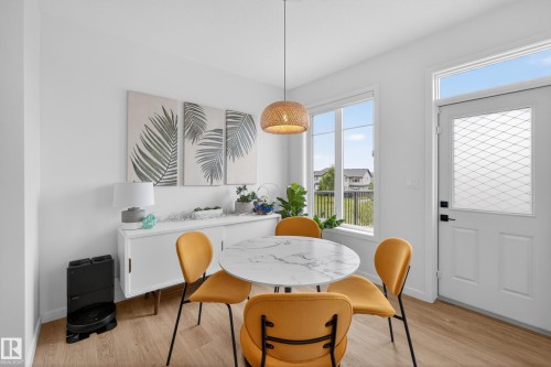 Inviting dining area featuring a round marble-finish table, wood-finish flooring, a contemporary pendant light fixture, a large window, and a white entry door with patterned glass insert and transom window - 17023 45 Street, Edmonton, AB 