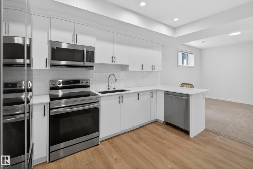 Modern kitchen featuring white cabinetry, stainless steel appliances, a subway tile backsplash, a dark basin sink, and wood-finish flooring - 17023 45 Street, Edmonton, AB 