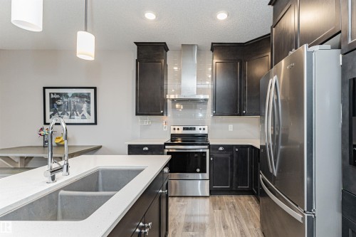 Modern kitchen featuring dark wood cabinetry, stainless steel appliances, a subway tile backsplash, light stone countertops, and wood-finish flooring - 8744 223 Street Nw, Edmonton, AB 
