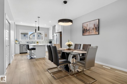 Open-concept living space featuring wood-finish flooring, a contemporary dining area with a circular pendant light fixture, and a kitchen with white cabinetry, stainless steel appliances, and a central island with pendant lighting - 1213 16A Street, Edmonton, AB 