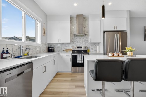 Contemporary kitchen featuring white cabinetry, stainless steel appliances, a white quartz countertop island with seating, and a marble-patterned backsplash - 1213 16A Street, Edmonton, AB 