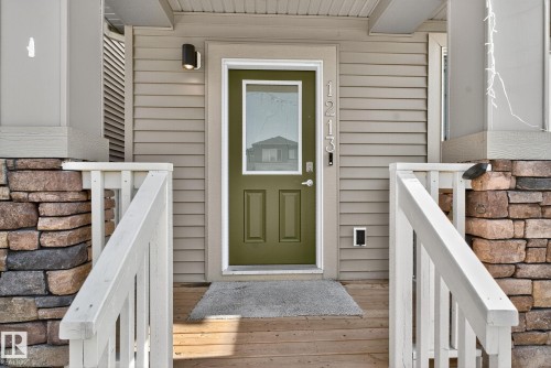 Entryway featuring a green paneled door with a clear glass insert, stone veneer columns, horizontal siding, and a wooden porch with white railings - 1213 16A Street, Edmonton, AB 