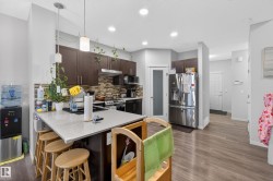Open-concept kitchen featuring dark wood-finish cabinetry, a light-toned countertop island with pendant lighting, a mosaic tile backsplash, stainless steel appliances, and wood-finish flooring - 
