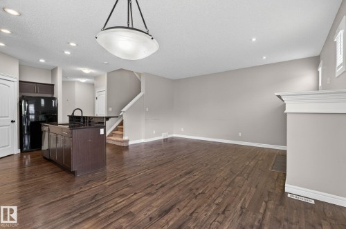 Open-concept living area featuring wood-finish flooring, a kitchen island with dark cabinetry and countertop, a black refrigerator, recessed lighting, and a fireplace with a white mantel - 64 South Creek Wynd, Stony Plain, AB 