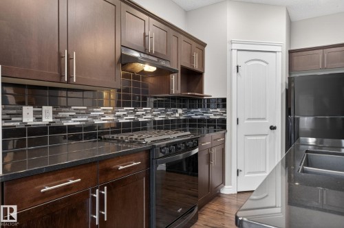 Kitchen featuring dark wood-finish cabinetry, black countertops, and a black tile backsplash with decorative accent tiles - 64 South Creek Wynd, Stony Plain, AB 