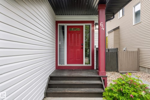 Entryway featuring a vibrant red door with glass side panels, a dark wood-finish porch, and white horizontal siding - 64 South Creek Wynd, Stony Plain, AB 