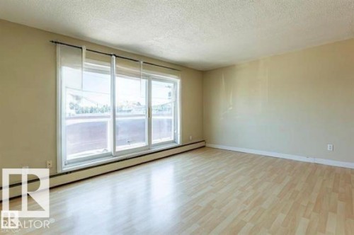 Spacious room with light wood-finish flooring, a large triple-panel window, and a perimeter baseboard heating unit - 307 8215 83 Avenue, Edmonton, AB 