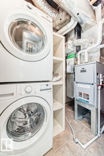 Dedicated laundry area featuring a stacked washer and dryer, adjacent built-in shelving, tile flooring, and visible utility systems - 4 11917 103 Street, Edmonton, AB 