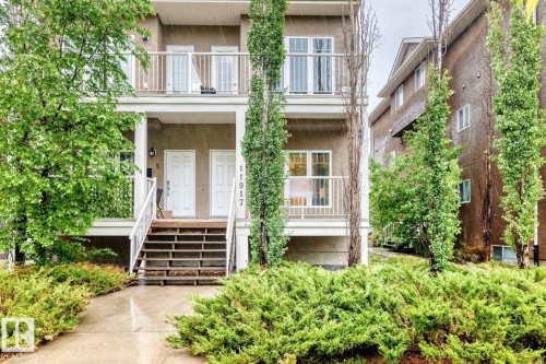 Multi-story residential facade featuring dual entry doors with overhead lighting, a second-story balcony with railings, and established landscaping with mature trees and shrubs - 4 11917 103 Street, Edmonton, AB 