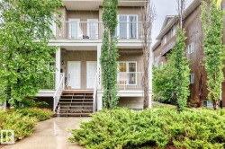 Multi-story residential facade featuring dual entry doors with overhead lighting, a second-story balcony with railings, and established landscaping with mature trees and shrubs - 