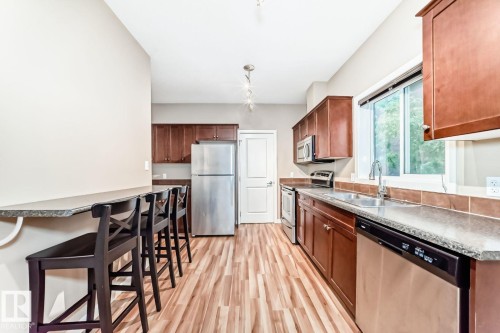 Modern kitchen featuring wood-finish flooring, stainless steel appliances, dark wood cabinetry, a built-in breakfast bar, and a window above the sink - 4 11917 103 Street, Edmonton, AB 