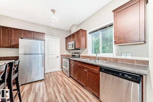 Kitchen featuring wood-finish flooring, stainless steel appliances, dark wood cabinetry, a double basin sink, and a window with natural light - 4 11917 103 Street, Edmonton, AB 