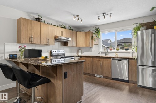 Open-concept kitchen featuring wood-finish cabinetry, granite-style countertops, stainless steel appliances, a kitchen island with seating, and wood-finish flooring - 33 Durrand Bend, Fort Saskatchewan, AB 