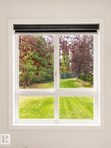 White framed window with a dark roller shade, offering a perspective of a grassy yard and mature trees - 115 5340 199 Street, Edmonton, AB - Indoor Photo Showing Other Room