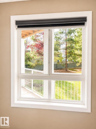 Double-paned window with bright white trim and integrated roller shades, offering a partial exterior patio deck with white railings and mature trees - 115 5340 199 Street, Edmonton, AB - Indoor Photo Showing Other Room