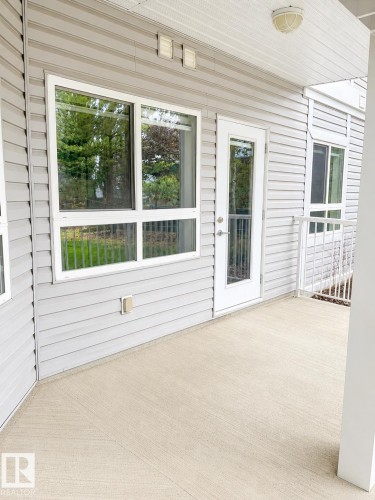 Covered concrete patio with textured finish, featuring light gray siding, multiple windows, a white framed door with glass paneling, and a white metal railing - 115 5340 199 Street, Edmonton, AB - Outdoor With Deck Patio Veranda With Exterior