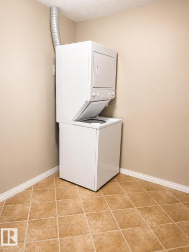 Dedicated laundry area featuring a stacked washer and dryer unit, neutral wall paint, white baseboards, and square tan floor tiles - 115 5340 199 Street, Edmonton, AB - Indoor Photo Showing Laundry Room