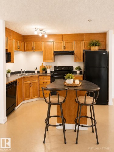 U-shaped kitchen with wood cabinetry, dark countertops, black appliances, and a central island with integrated seating - 115 5340 199 Street, Edmonton, AB - Indoor Photo Showing Kitchen With Double Sink