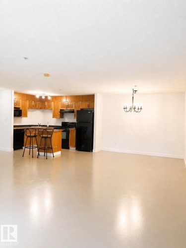 Open-concept kitchen and living area featuring a breakfast bar with seating, black appliances, warm wood cabinetry, and a black chandelier - 115 5340 199 Street, Edmonton, AB - Indoor Photo Showing Other Room