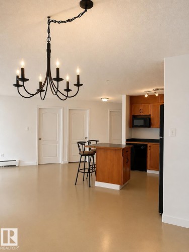 Spacious open-concept living area featuring a black chandelier, light-toned flooring, white walls, and a kitchen with wood-finish cabinetry, black countertops, and black appliances - 115 5340 199 Street, Edmonton, AB - Indoor