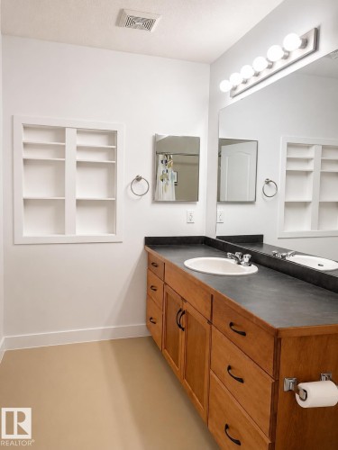 Bathroom featuring a wood-finish vanity with an integrated sink, dark countertop, and large mirror with globe lighting - 115 5340 199 Street, Edmonton, AB - Indoor Photo Showing Bathroom