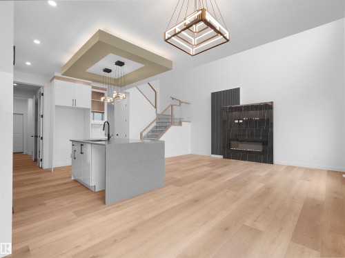 Open-concept living area featuring wide plank wood-finish flooring, a contemporary kitchen island with an undermount sink, a recessed ceiling with integrated lighting, a modern fireplace with a dark stone surround, and a staircase with glass railings - 214 Crystal Creek Drive, Leduc, AB 