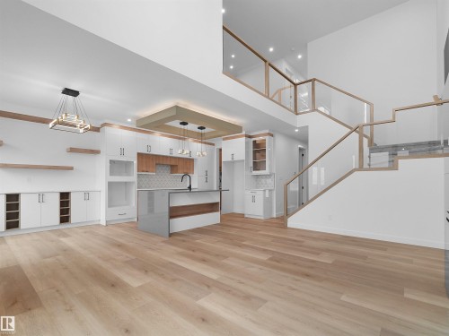 Expansive open-concept interior featuring light wood-finish flooring, a contemporary kitchen island with a dark faucet, white cabinetry with wood accents, and a multi-level staircase with glass railings - 214 Crystal Creek Drive, Leduc, AB 