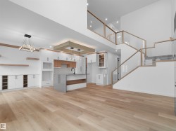 Expansive open-concept interior featuring light wood-finish flooring, a contemporary kitchen island with a dark faucet, white cabinetry with wood accents, and a multi-level staircase with glass railings - 