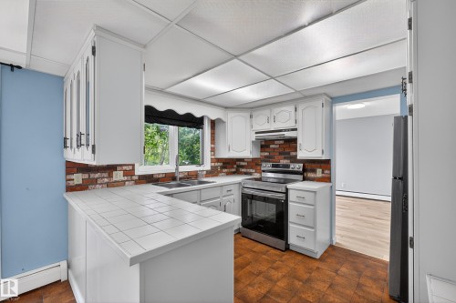 Kitchen featuring white cabinetry, a brick-style backsplash, tile countertops, stainless steel appliances, and wood-finish flooring - 14808 55 Street, Edmonton, AB 