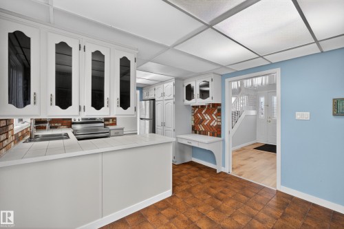 Kitchen featuring white cabinetry with glass inserts, tile countertops, and a brick-patterned backsplash - 14808 55 Street, Edmonton, AB 