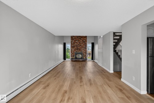 Spacious living area featuring wood-finish flooring, a prominent brick-clad fireplace with a dark metal insert, and light gray wall paint - 14808 55 Street, Edmonton, AB 