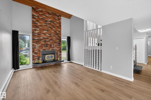 Vaulted ceiling with exposed wood beam, brick fireplace with wood-burning insert and slate hearth, light wood-finish flooring, white baseboards, and staircase with painted wood railing - 14808 55 Street, Edmonton, AB 