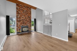 Vaulted ceiling with exposed wood beam, brick fireplace with wood-burning insert and slate hearth, light wood-finish flooring, white baseboards, and staircase with painted wood railing - 