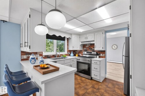 Bright kitchen featuring white cabinetry, a tiled breakfast bar, brick-look backsplash, and stainless steel appliances including a gas range - 14808 55 Street, Edmonton, AB 
